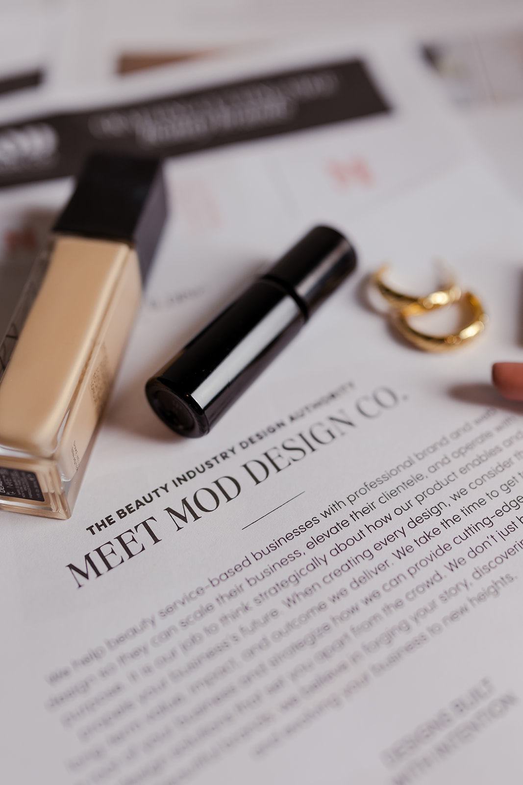 A close-up image showing a beauty product flat lay with a bottle of foundation, a black lip product, and a pair of gold hoop earrings placed on top of a printed document. The document's headline reads "Meet MOD Design Co." and below it, a description discusses how MOD Design Co. helps beauty-based businesses elevate their clientele through strategic branding and design. The design layout is stylish and professional, in line with the beauty industry's high-end aesthetic.